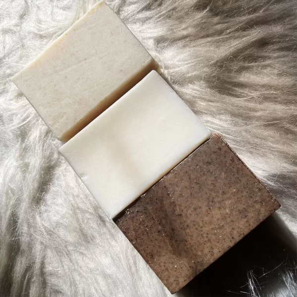 Three bars of Coconut Oil Soap with Gentle Exfoliating Cacao Shells, placed on a fluffy, white fur surface. the top bar is plain, the middle is creamy, and the bottom has an exfoliating texture.