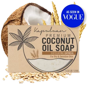 A box of Coconut Oil Soap with Exfoliating Oatmeal features an illustration of a coconut palm tree and text indicating exfoliating oatmeal for dry and sensitive skin. The box is placed in front of a split coconut, wheat stalks, and scattered oats. A blue "As Seen in Vogue" label is in the corner.