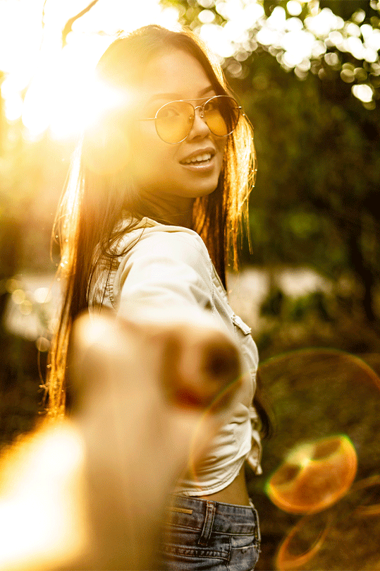 A joyous woman reaching out to the camera with a beaming smile, bathed in the golden glow of sunset light filtering through trees, her hand grasping a coconut.