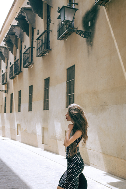 A woman in a coconut polka dot dress strolling thoughtfully through a sunlit European alleyway, her hair gently tousled by the breeze.
