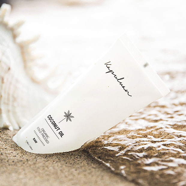 A tube of coconut oil product resting on a sandy beach with a coconut shell nearby, as gentle waves lap at the shore.