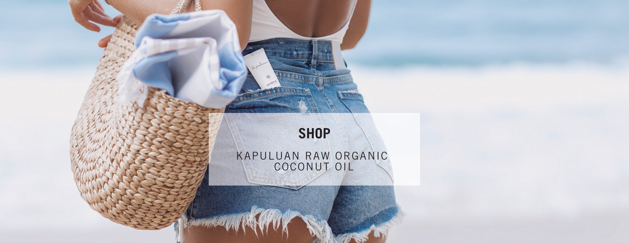 A person holding a coconut straw tote bag with a partial view of a kapuluan raw organic coconut oil product, dressed in denim shorts and standing against a blurred beach backdrop.