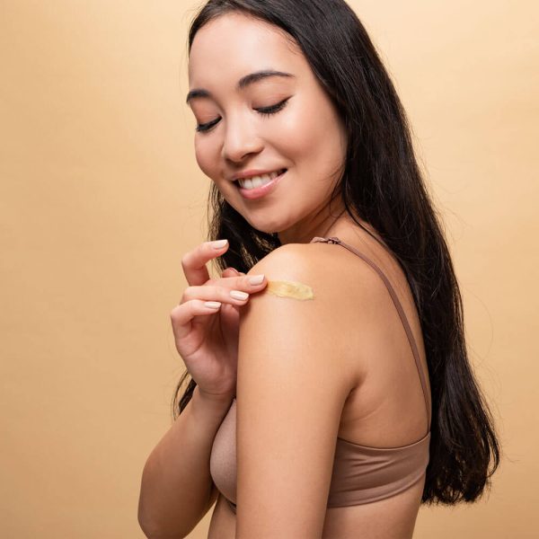 A woman applying a patch of Organic Cocoa Butter skin-tone bandage to her upper arm, with a content expression on her face against a beige background.