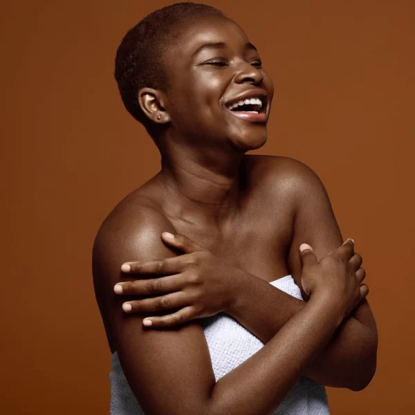 A smiling person with short hair is wrapped in a white towel against a solid brown background. They are crossing their arms across their chest, exuding happiness and confidence after using Muscle Relief Coconut Body Oil.