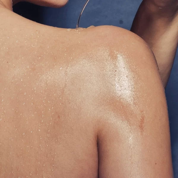 Close-up of a person's wet shoulder and upper back, glistening with water droplets against a dark background. The image captures the smooth texture of the skin and the play of light on the water, highlighting the moisture and subtle contours of the shoulder, enhanced by Muscle Relief Coconut Body Oil.