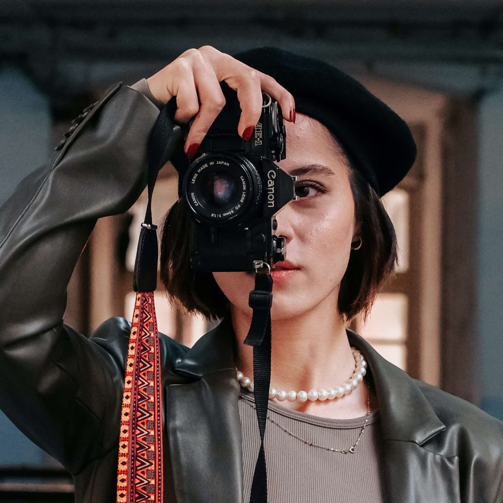 A young woman with a beret and pearl necklace holds a canon camera to her face in a focused pose, ready to snap a photo. her expression is intense and concentrated.