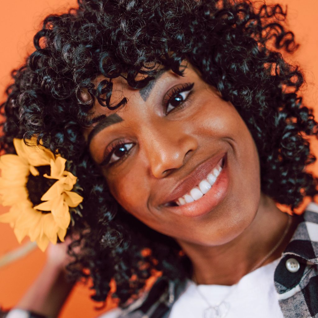 A smiling woman with curly hair and a yellow flower tucked behind her ear, against a warm orange background.