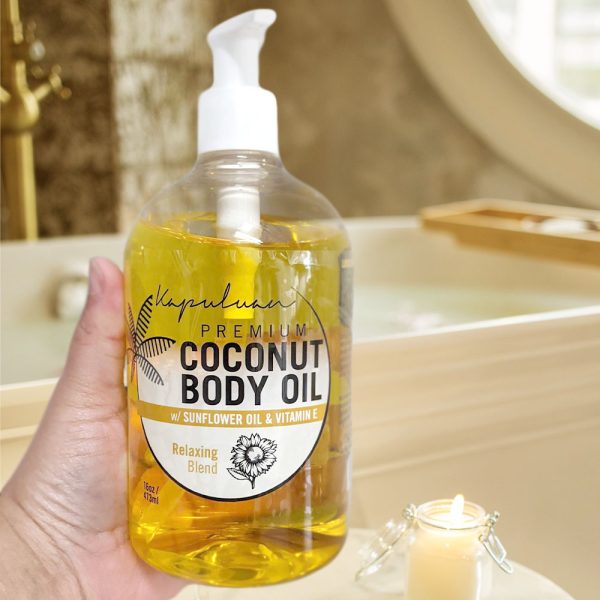 A hand holds a transparent bottle of Relaxing Coconut Body Oil with a label featuring sunflower oil and vitamin e, in a serene bathroom setting with a lit candle in the background.