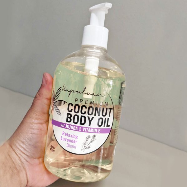 A hand holding a bottle of Relaxing Coconut Body Oil. The bottle features a pump dispenser and a label indicating it contains jojoba, vitamin E, and a relaxing lavender blend. The background is a plain, neutral color.