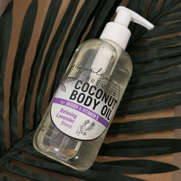 A bottle of Relaxing Coconut Body Oil with jojoba and vitamin E, featuring a relaxing lavender blend, rests on green palm leaves. The bottle is equipped with a pump dispenser.