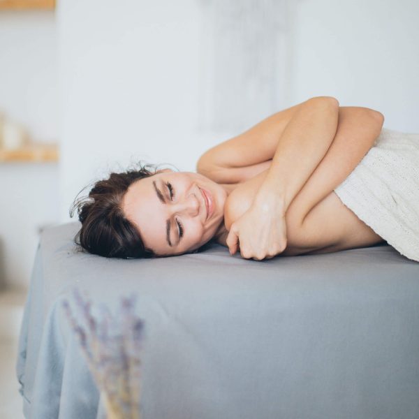 A person resting on a massage table, smiling blissfully with eyes closed and enjoying a moment of relaxation, enveloped in the gentle scent of Relaxing Coconut Body Oil.