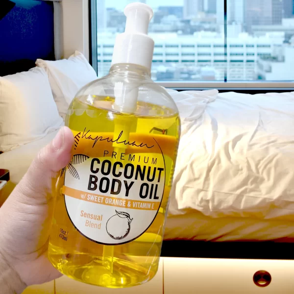 Close-up of a hand holding a pump bottle of Sensual Coconut Body Oil. The background shows an unmade bed by a window with an urban cityscape visible outside.