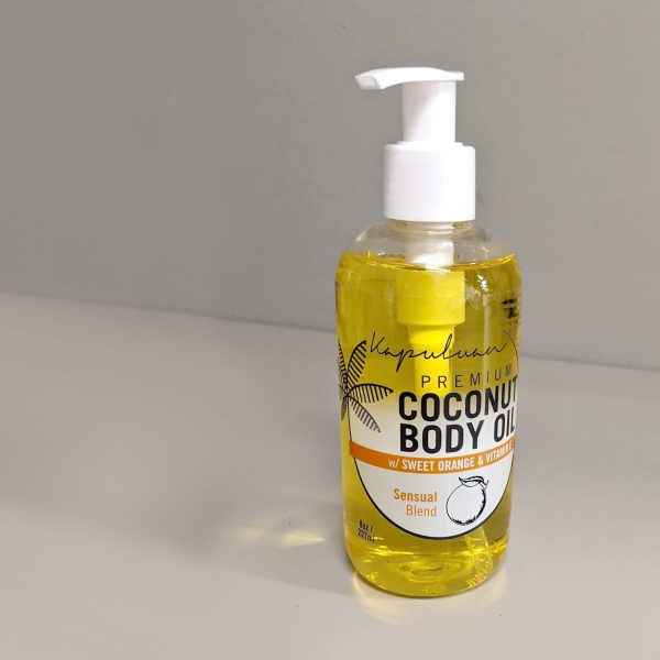 A bottle of Sensual Coconut Body Oil with sweet orange and vanilla on a gray countertop, against a light background. the oil is golden yellow and the pump dispenser is white.