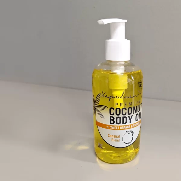A bottle of Sensual Coconut Body Oil sits on a white surface. The bottle has a white pump dispenser and a label featuring palm tree leaves. The product is described as a "Sensual Blend".