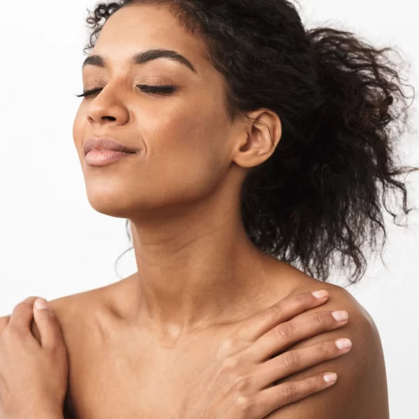 A person with curly hair pulled back smiles gently with closed eyes, appearing relaxed and serene. They are topless with hands gently resting on their shoulders, showcasing the Sensual Coconut Body Oil against a plain white background.