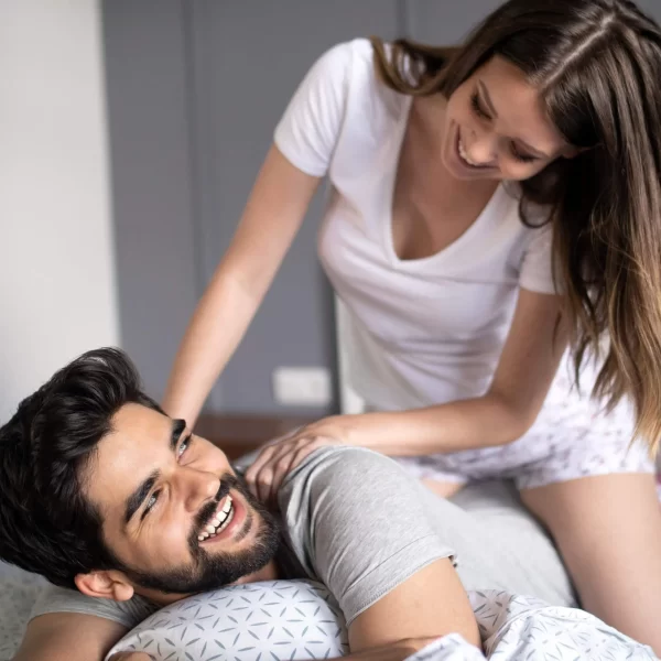 A smiling man with a beard is lying down on a bed, resting his head on a pillow. A woman with long brown hair, wearing a white shirt and patterned shorts, leans over him, touching his shoulder and smiling at him. They look happy and engaged with each other as they enjoy the Sensual Coconut Body Oil.