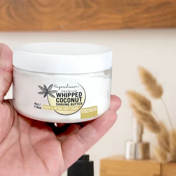 A hand holds a jar of Whipped Coconut Shaving Butter. The label states it provides a silky and ultra-soft shave. The jar has a black palm tree logo. In the background, there is a blurred wooden object and dried floral arrangements.