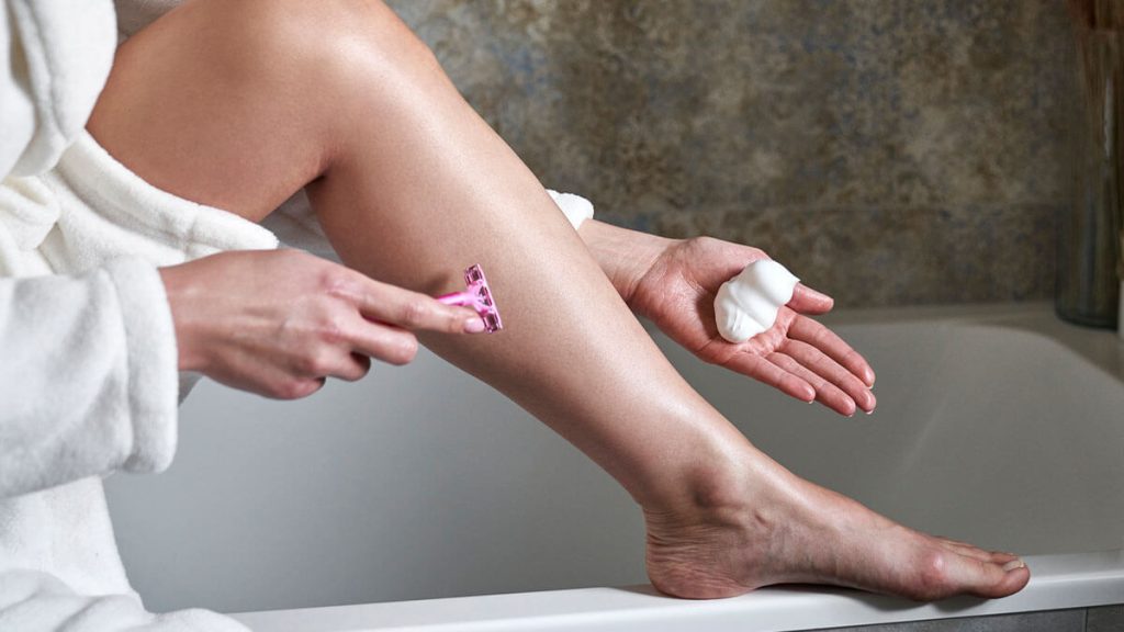 A person in a white robe is applying Whipped Coconut Shaving Butter on their leg while holding a pink razor, seated beside a bathtub.