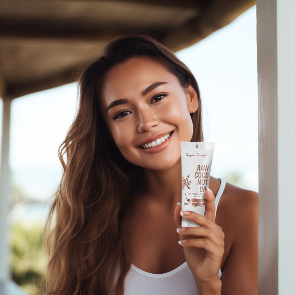 A smiling young woman holding a bottle of coconut oil skincare product, standing under a shaded porch with greenery and sunlight in the background.