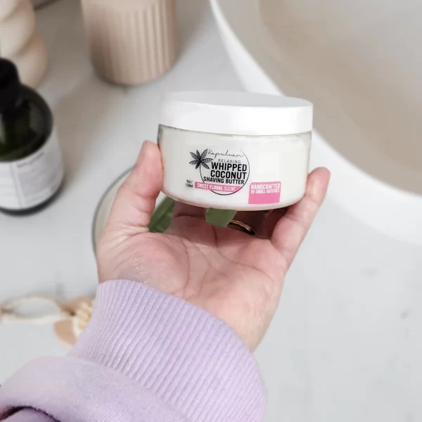 A person in a lavender sweater sleeve is holding a jar of Whipped Coconut Shaving Butter. The jar is white with a black lid and a pink label. Blurred background shows a bathroom countertop with various toiletries.