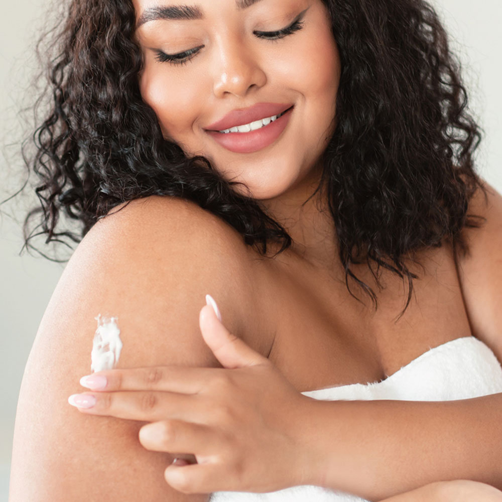 A person with wavy hair is smiling and applying a dollop of cream onto their shoulder. They are wrapped in a white towel, suggesting they might be getting out of or preparing for a bath or shower.