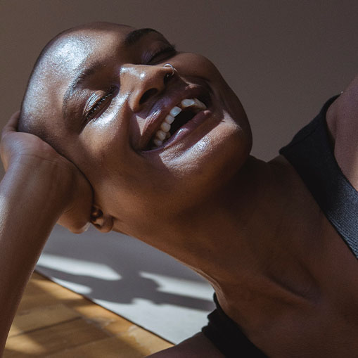 A joyful black woman with a shaved head, laughing and holding her head with one hand, bathed in natural sunlight.
