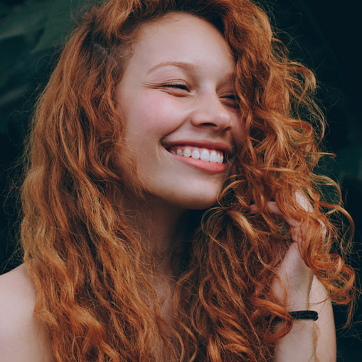 A joyful woman with curly red hair smiling broadly, her eyes are gently closed, and the background is softly blurred.