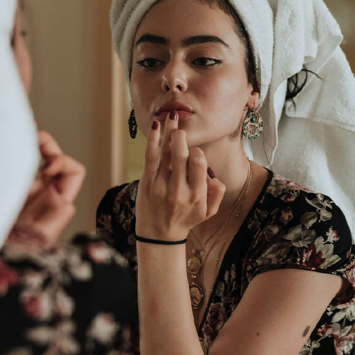 A woman with a towel wrapped around her hair applies lipstick in front of a mirror. she's wearing a floral dress and has decorative earrings, focusing intently on her reflection.