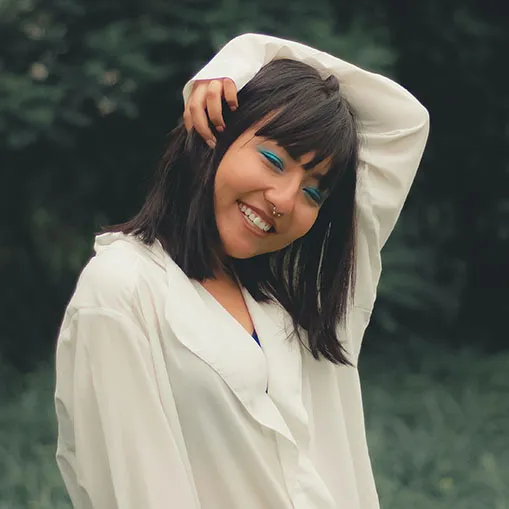 A person with a nose ring and bright blue eye makeup is smiling and posing outdoors. They have shoulder-length dark hair with bangs and are wearing a loose white blouse. One hand is touching their head, creating a relaxed and happy demeanor.