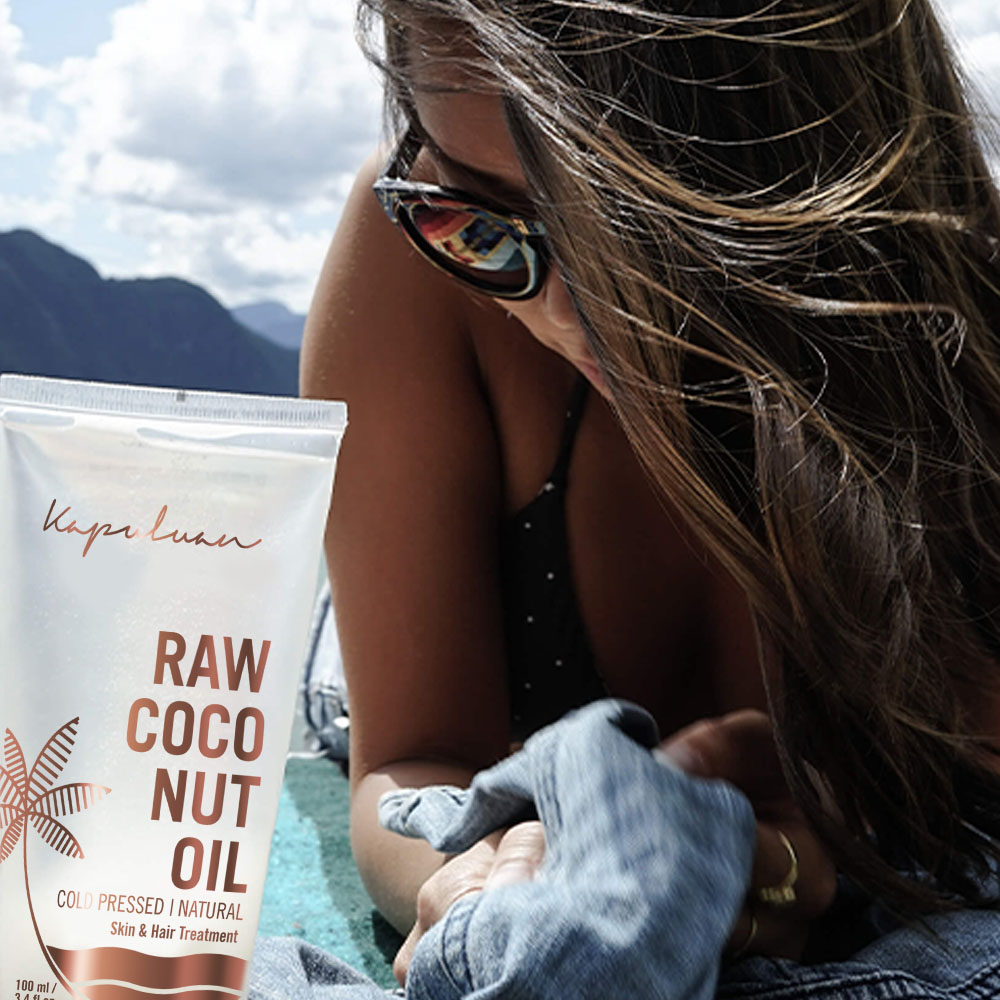 A woman in sunglasses reclines by a body of water, holding a transparent pouch of "raw coconut oil." her focus is on the product, which is prominently displayed in the foreground.