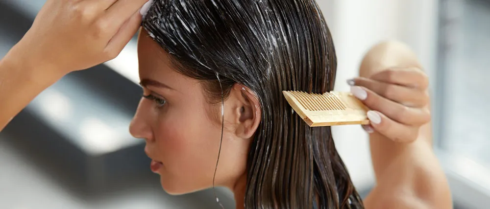 A person combs conditioner through their wet, dark hair with a wooden comb. Their face is partially visible, and they are focusing on their hair care routine. The background is blurred.