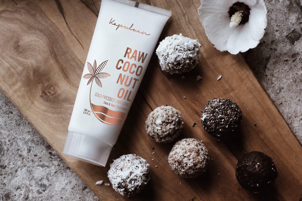 Tube of raw coco nut oil on a wooden surface, surrounded by coconut-dusted truffles, with a white flower nearby, emphasizing natural skin care and indulgence.