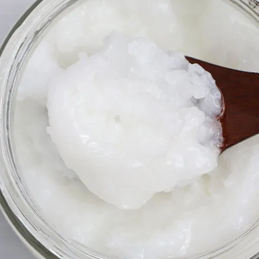 A close-up view of a glass jar filled with white coconut oil. The texture appears semi-solid, and a wooden spoon is scooping out some of the oil from the jar. The oil has a smooth and creamy consistency.