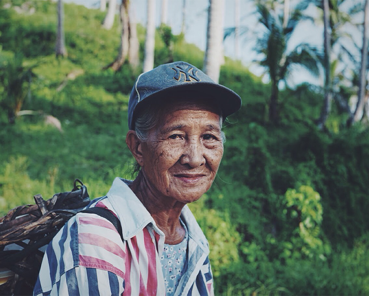 An elderly woman wearing a baseball cap and a striped shirt, smiling gently. she is outdoors with lush greenery and palm trees in the background.