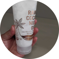 A hand holding a tube of raw coconut oil skincare product, designed for both skin and hair use, with labels emphasizing its cold-pressed qualities.