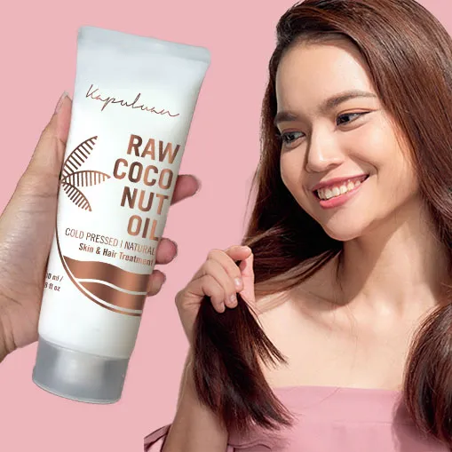 A hand is holding a tube of Kapuluan Raw Coconut Oil against a pink background. Next to the product, a woman with long, straight hair smiles while touching her hair. The text on the tube highlights that it is cold-pressed and natural for skin and hair treatment.