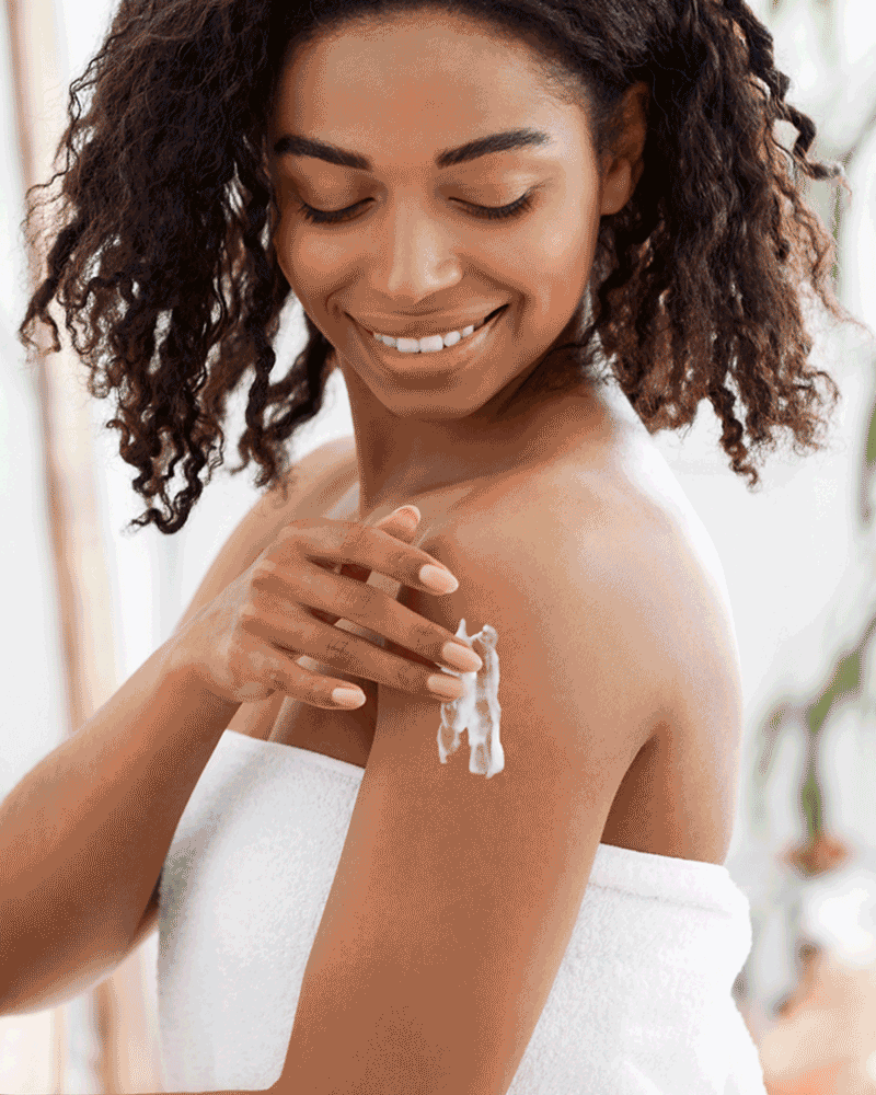 A woman with a radiant smile applies coconut oil lotion to her shoulder, standing against a bright, blurred background. Her curls cascade around her face, enhancing her joyful expression.