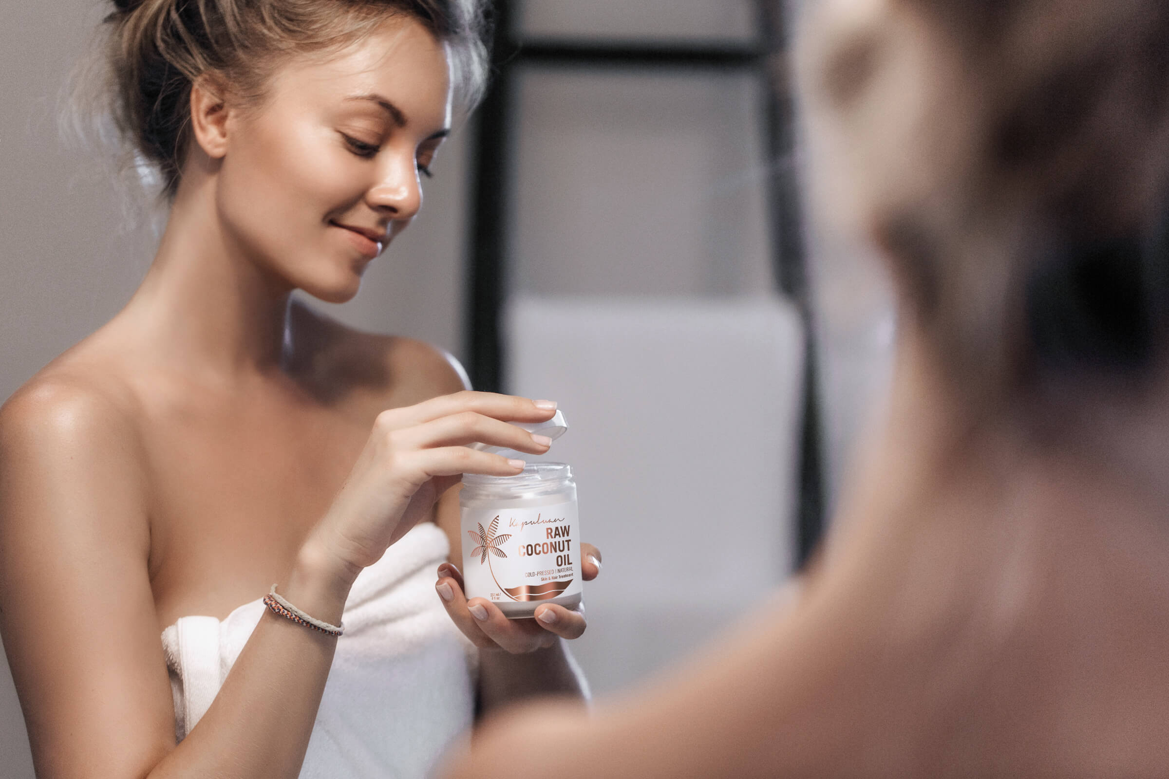 A woman wearing a towel looks at a jar labeled 'Cold-Pressed Raw Coconut Oil' in her hand, smiling as she stands before a mirror in a well-lit bathroom.