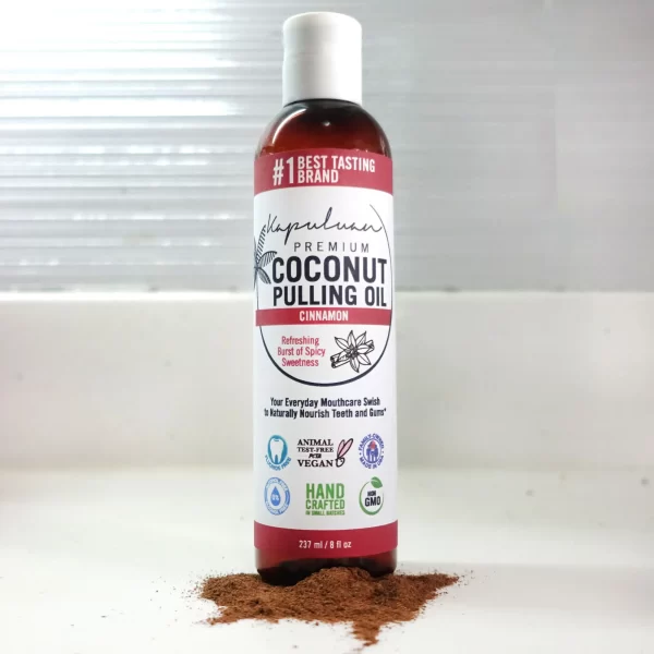 A bottle of Coconut Pulling Oil - Cinnamon is placed on a white surface with a small pile of cinnamon powder at the base. The label highlights features like non-GMO, vegan, animal cruelty-free, and handcrafted. The bottle contains 237ml (8 fl oz).