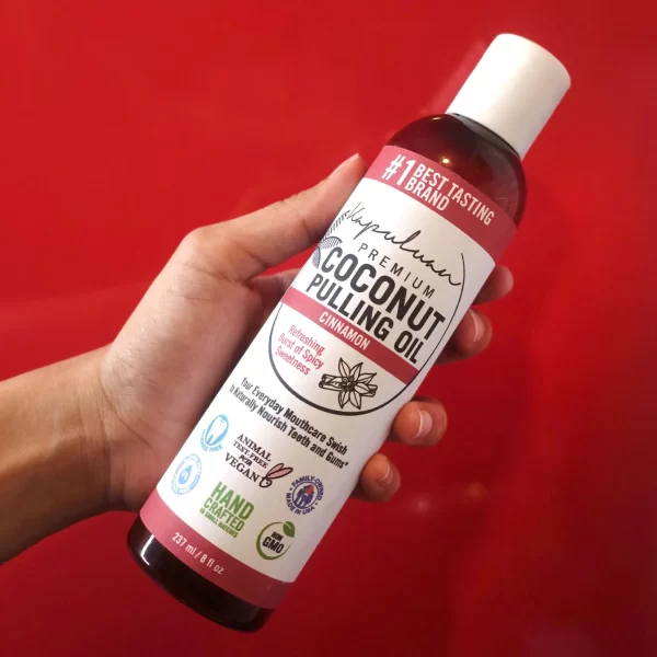 A hand holds a bottle of Coconut Pulling Oil - Cinnamon. The label highlights that it is vegan, non-GMO, and good for everyday mouthcare and swish. The bottle is tall and white with red accents, against a red background.