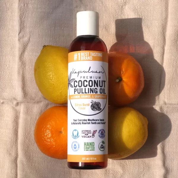 A bottle of Coconut Pulling Oil - Citrus Burst with lemon, orange, and grapefruit flavor is placed on a cloth with two lemons and two oranges surrounding it. The bottle features various labels, including best tasting brand, non-GMO, and handcrafted.