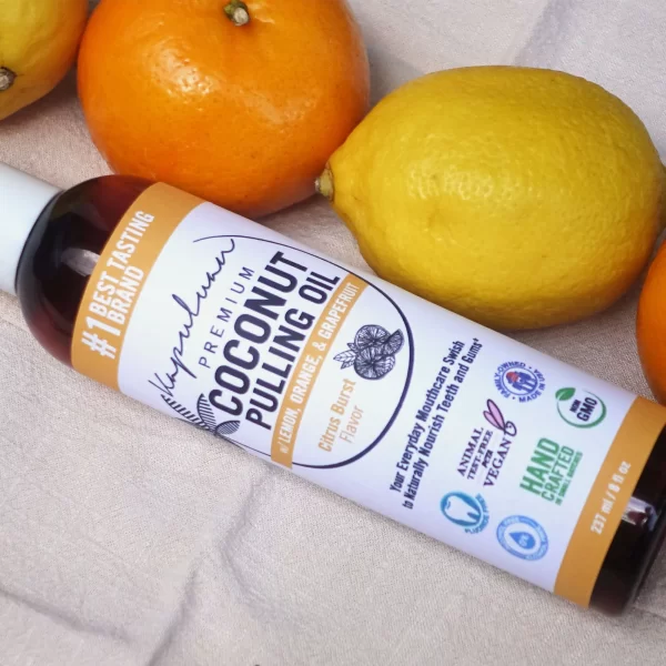 A bottle of coconut pulling oil is placed on a light fabric surface, surrounded by lemons and oranges. The label on the bottle reads "Coconut Pulling Oil - Citrus Burst" and highlights the lemon, orange, and grapefruit flavor. Various certifications are also visible on the label.