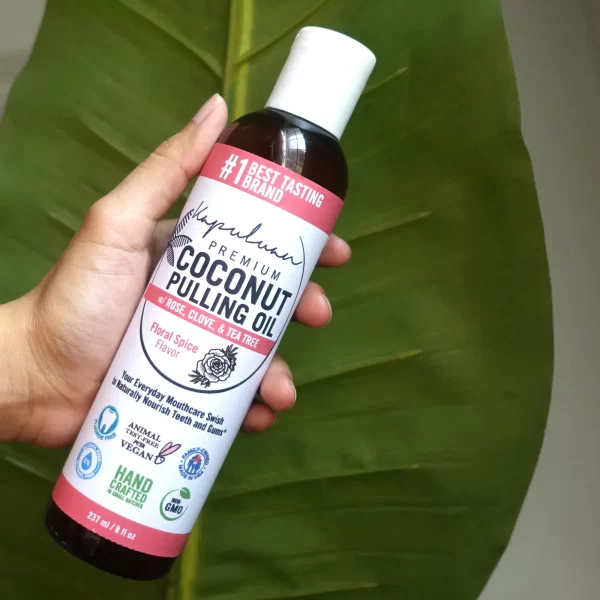 Hand holding a bottle of Coconut Pulling Oil - Floral Spice against a green leaf background. The label indicates it includes rose, clove, and tea tree, with a floral spice flavor. The product is touted as a chemical-free, hand-crafted, and vegan-friendly oral care.