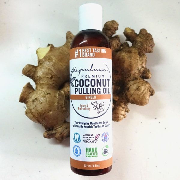 A bottle of Coconut Pulling Oil - Ginger is standing upright on a tabletop, surrounded by fresh ginger roots. The label highlights it as handcrafted, non-GMO, vegan, and designed for oral care.