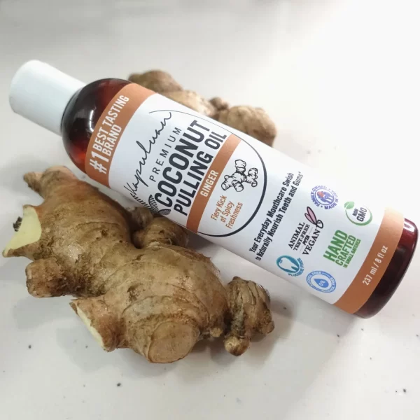 A bottle labeled "Coconut Pulling Oil - Ginger" lies on a white surface next to a piece of fresh ginger root. The bottle has various certifications, including Non-GMO, Vegan, and Handcrafted with a volume of 237ml.