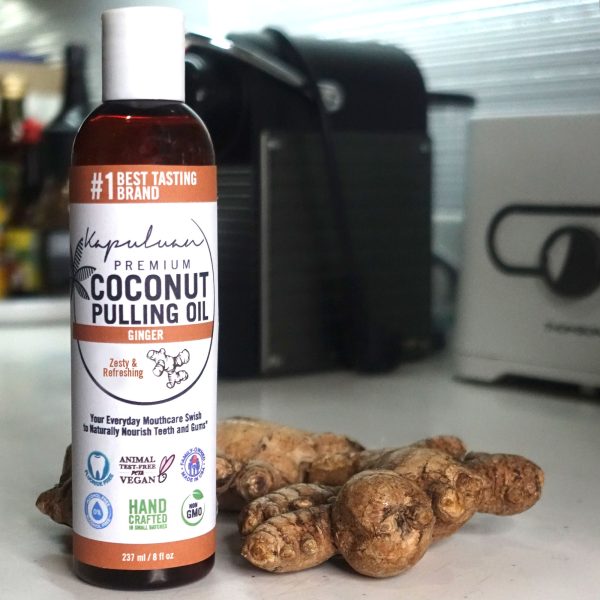 A bottle of Coconut Pulling Oil - Ginger resting on a kitchen counter beside fresh ginger roots. The label emphasizes its zesty and refreshing qualities, proudly marking it as a vegan, non-GMO, handcrafted product.