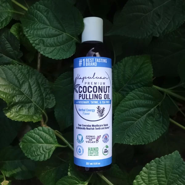 A bottle of Coconut Pulling Oil - Herbal Energy is placed against a backdrop of green leaves. The label highlights attributes such as rosemary, thyme, tea tree, being animal cruelty-free, GMO-free, and handcrafted.