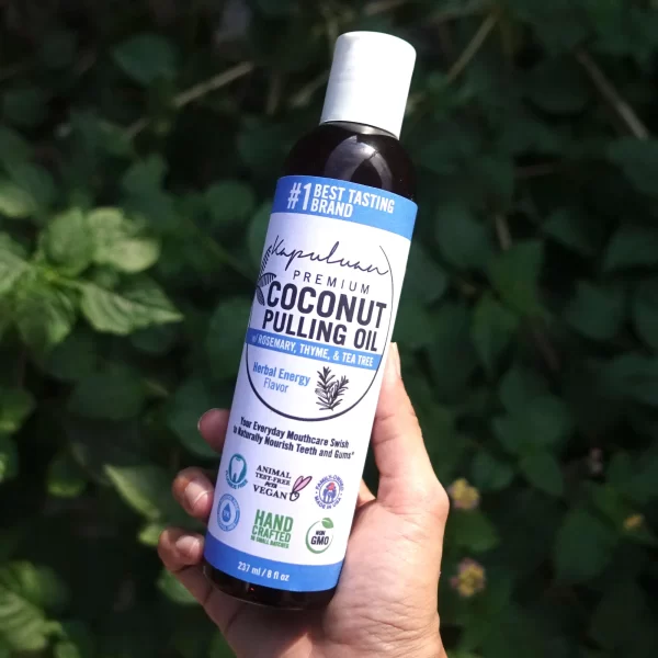 A hand holding a bottle of Coconut Pulling Oil - Herbal Energy against a leafy background. The label features details like "Rosemary, Thyme & Tea Tree," "Herbal Energy flavor," "Animal Test Free & Vegan," "Non-GMO," and "Hand-Crafted in Small Batches.