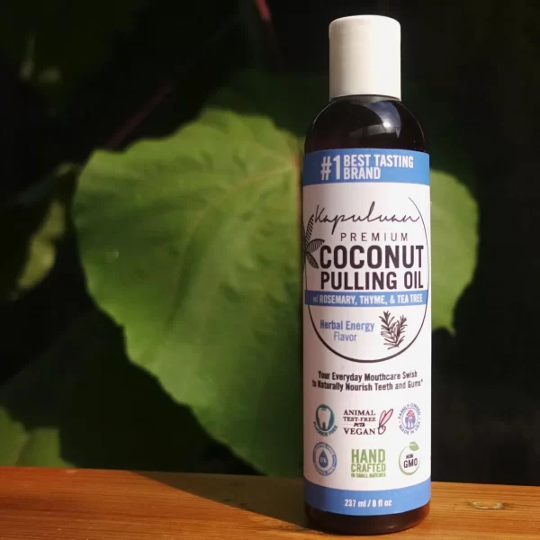 A bottle of Coconut Pulling Oil - Herbal Energy, with "Rosemary, Thyme, & Tea Tree" and "Herbal Energy Flavor" on the label. It promotes dental health, is handcrafted, non-GMO, animal and cruelty-free, and suitable for vegans. The bottle is set against a leafy background.