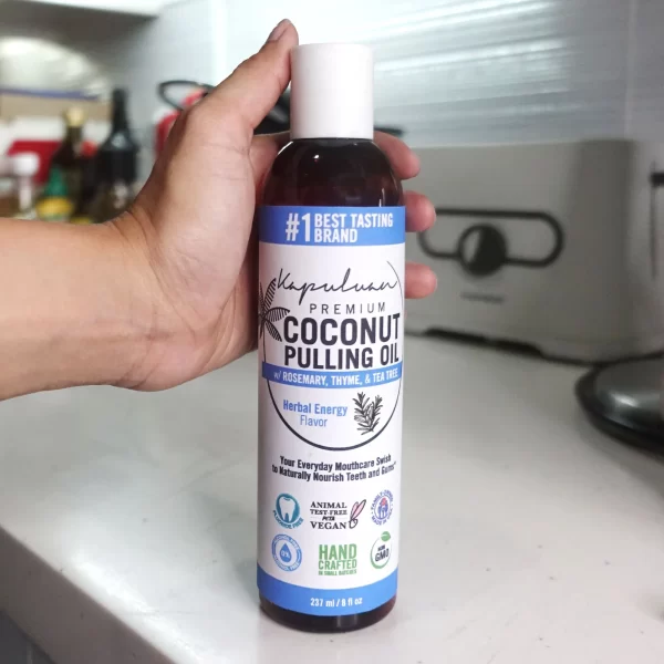 A hand is holding a dark brown bottle of Coconut Pulling Oil - Herbal Energy in a kitchen. The bottle has a white label with blue, green, and black text and icons. It features "Best Tasting Brand," "Kapuluan," and mentions rosemary, thyme, and tea tree.